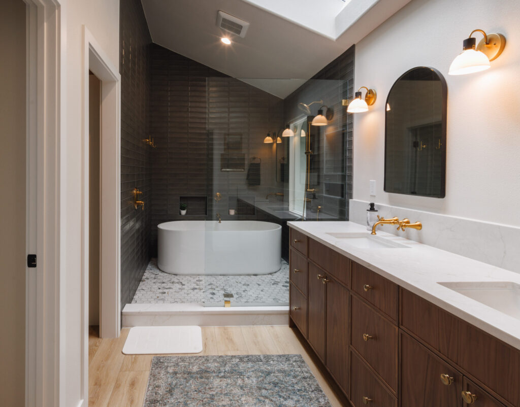 A beautiful primary bathroom remodel featuring a large wet room with a shower and a freestanding tub. There are natural wood cabinets, quartz countertops and beautiful brushed gold faucets and vanity lights.